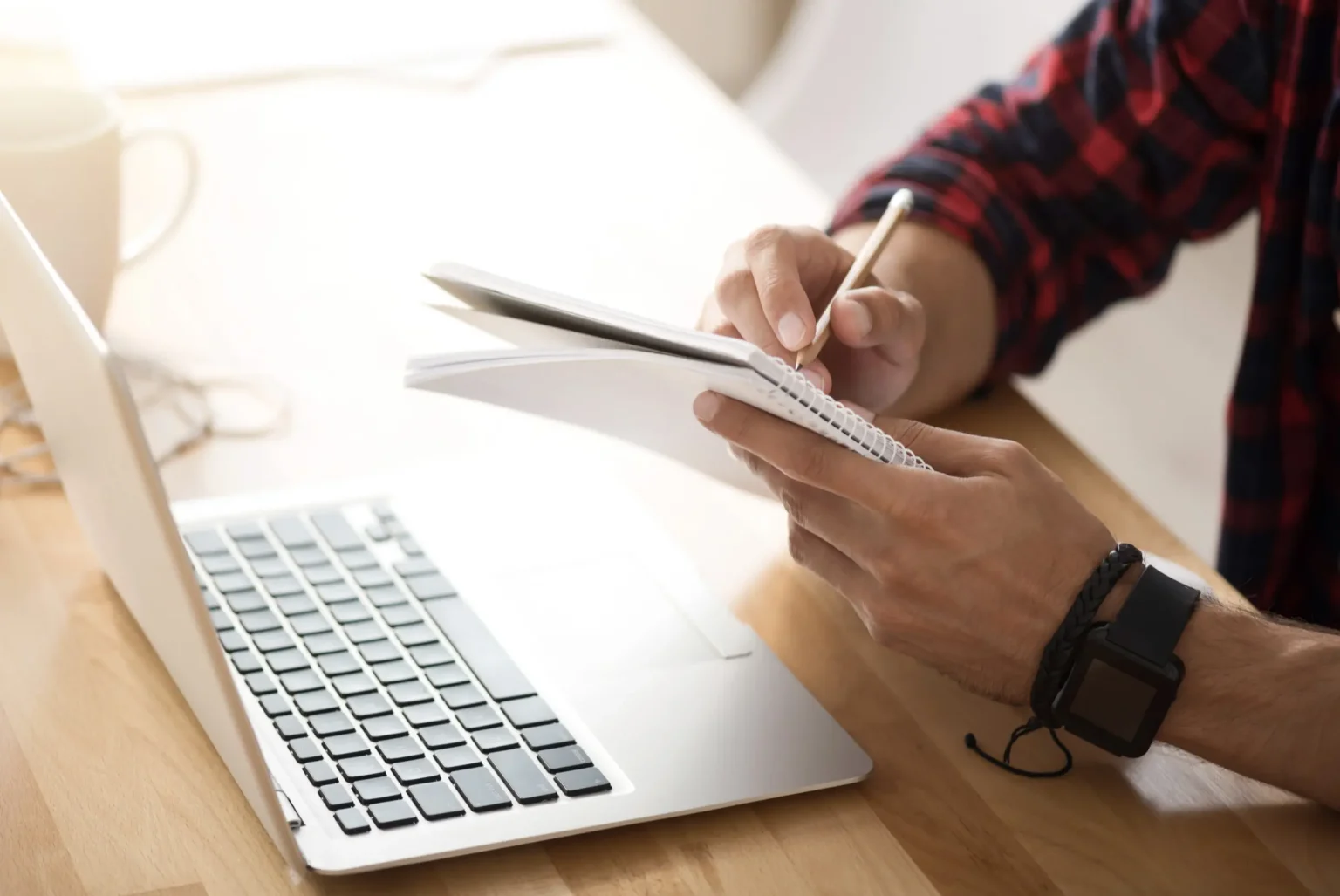 Close-up of a person's hands writing in a spiral notebook beside an open laptop on a wooden desk, warm neutral tones, calm and focused mood